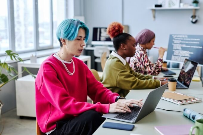 Tim beragam pengembang perangkat lunak Gen Z yang bekerja di kantor, dengan fokus pada seorang pemuda muda Asia dengan rambut berwarna menggunakan komputer