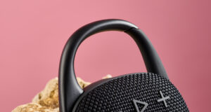 Close up dari carabiner di atas JBL Clip 5, digambarkan bersandar pada batu berwarna pasir dengan latar belakang merah muda,