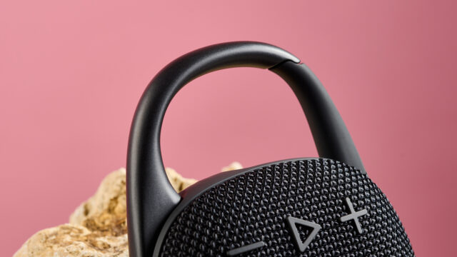 Close up dari carabiner di atas JBL Clip 5, digambarkan bersandar pada batu berwarna pasir dengan latar belakang merah muda,