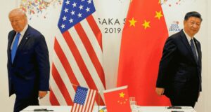 Mantan Presiden Donald Trump berpidato di podium dengan ekspresi tegas, bendera AS dan Tiongkok dipajang berdampingan di latar belakang.